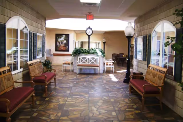 Indoor common-area corridor with benches, potted plants, a central clock and decorative lamp posts.