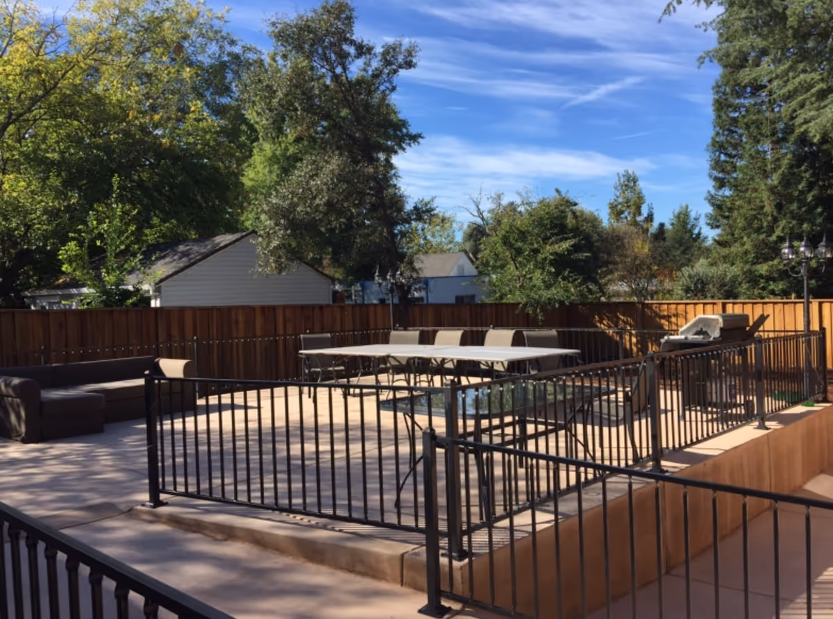 Outdoor patio area with a black metal fence surrounding a seating area that includes a brown outdoor sofa, a table with several chairs, and a barbecue grill. Trees and a wooden fence are visible in the background under a blue sky with some clouds.