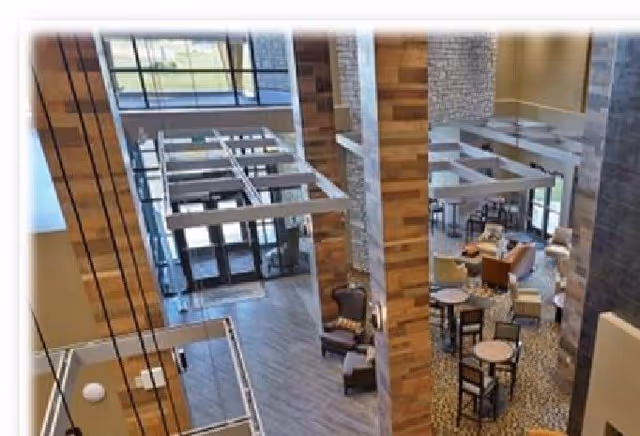 Interior view of a senior living facility common area with high ceilings, large windows, wooden pillars, and multiple seating arrangements including chairs and tables.