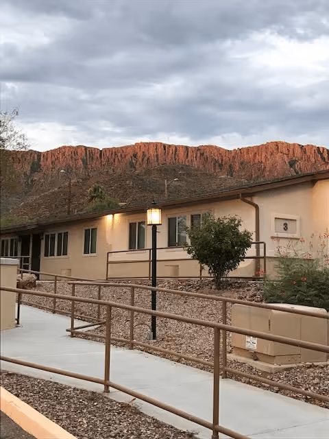 Exterior view of a single-story building with beige walls and multiple windows, a walkway with handrails, a lamp post, and desert landscaping with rocks and shrubs. In the background, there is a red rocky mountain under a cloudy sky.