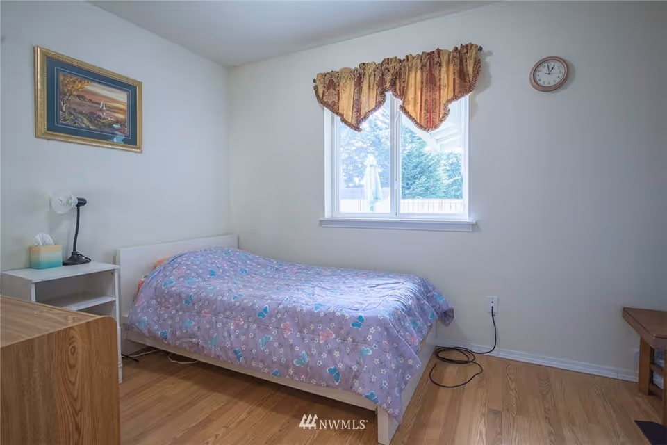 A small bedroom with a single bed covered in a purple blanket with butterfly patterns. There is a white nightstand with a lamp and tissue box next to the bed. A framed picture hangs on the wall above the nightstand. A window with patterned curtains lets in natural light, and a clock is mounted on the wall to the right of the window. The floor is wooden, and some electrical cords are visible near the baseboard.