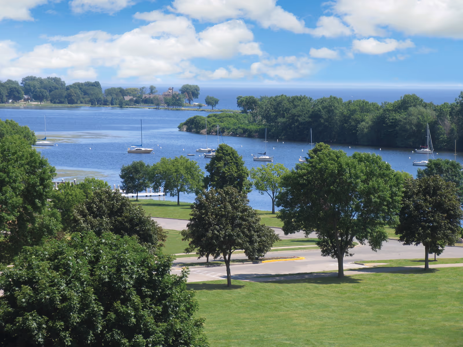 View of a serene waterfront with several sailboats anchored near the shore, surrounded by lush green trees and a grassy area with a road running through it under a partly cloudy blue sky.