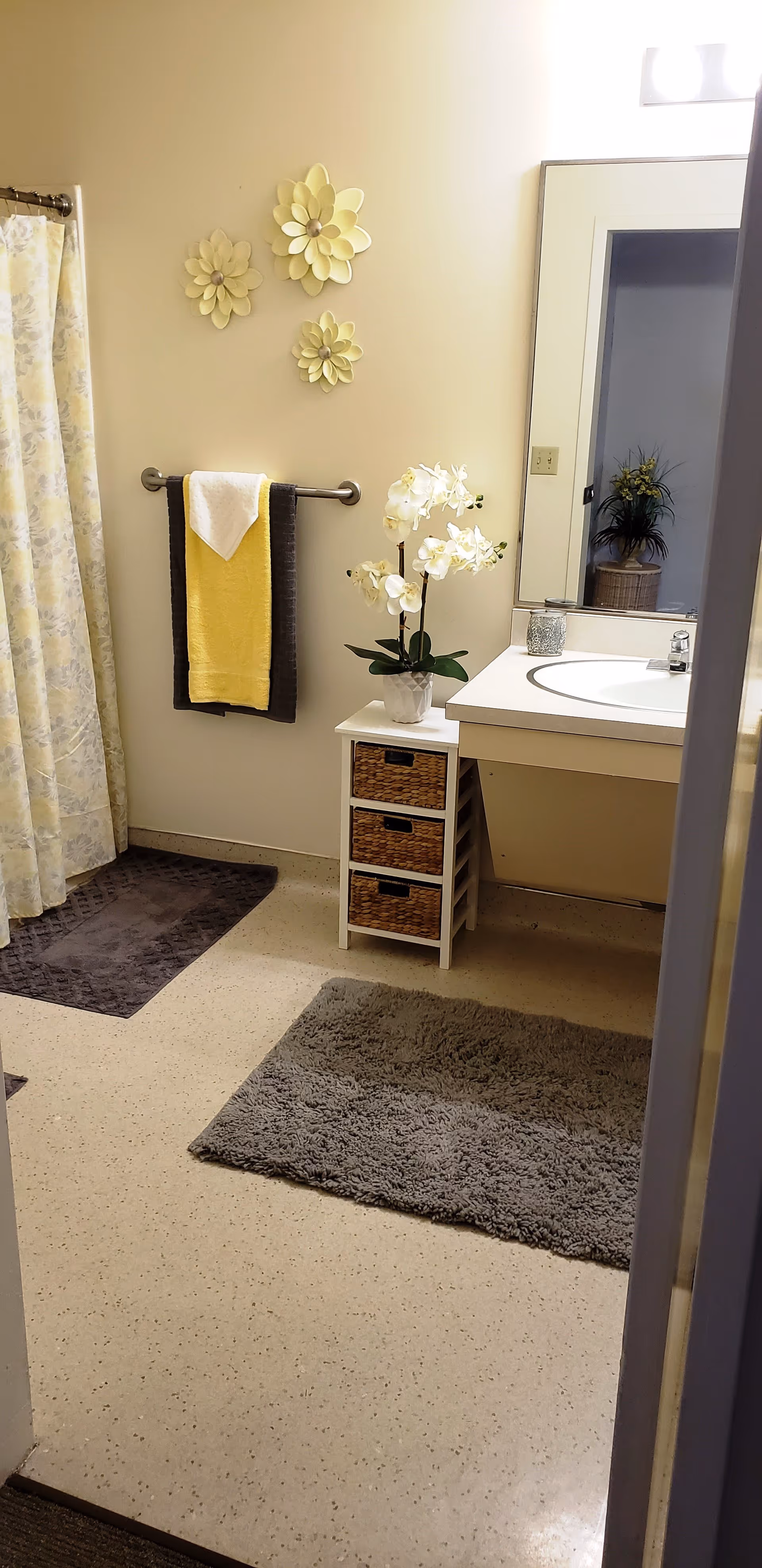 A clean bathroom with a shower curtain on the left, two dark gray bath mats on the floor, a towel rack with a yellow and white towel, three decorative yellow flower wall hangings, a white vanity with a sink, a small cabinet with wicker drawers, and a white orchid plant on top. A large mirror is mounted above the sink reflecting a plant in the adjacent room.