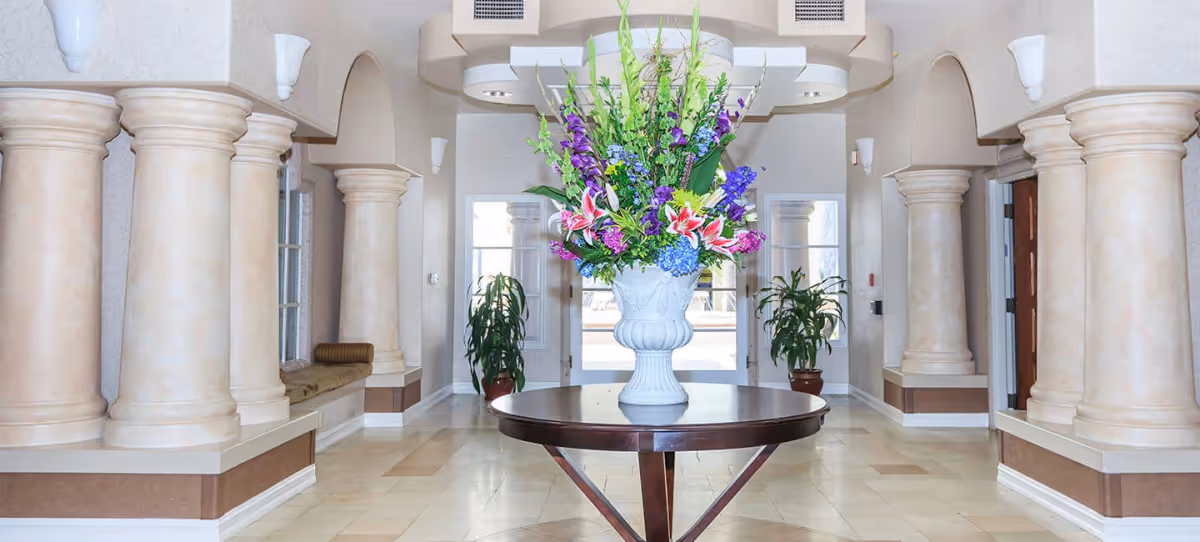 A spacious and elegant interior hallway with large beige columns on both sides, a round wooden table in the center holding a large white vase filled with colorful flowers including purple, pink, and blue blooms. There are two potted plants near the glass doors at the far end, and the walls are light-colored with arched openings and sconces.