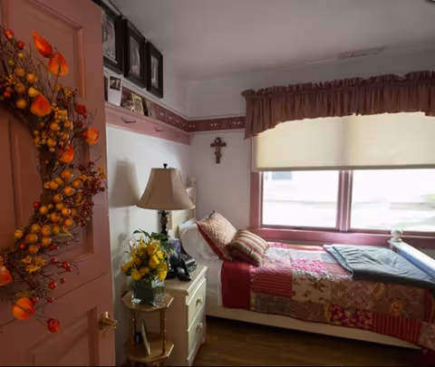 A cozy bedroom with a single bed covered in a colorful patchwork quilt and several pillows. Next to the bed is a white nightstand with a lamp, a telephone, and a vase of yellow flowers. A window with a brown valance and a closed blind lets in natural light. The door is decorated with a fall-themed wreath, and a cross hangs on the wall above the bed.
