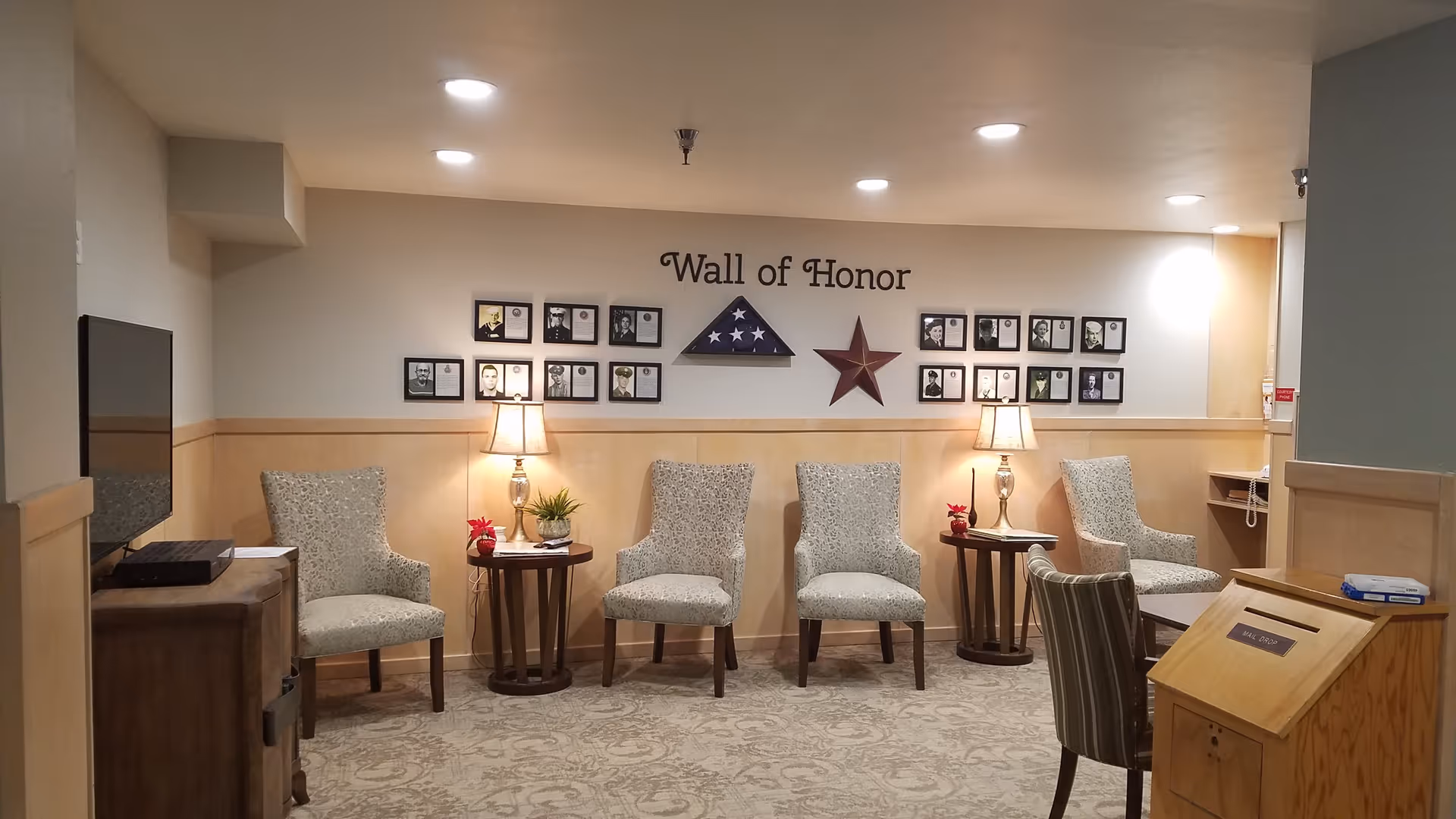 A cozy seating area in a senior living facility with four patterned armchairs arranged along a wall. Two small round tables with lamps and small plants are placed between the chairs. Above the chairs, a 'Wall of Honor' displays framed photos and a folded American flag in a triangular case, along with a decorative star. A TV is mounted on the left side, and a wooden mail drop box is visible on the right side.