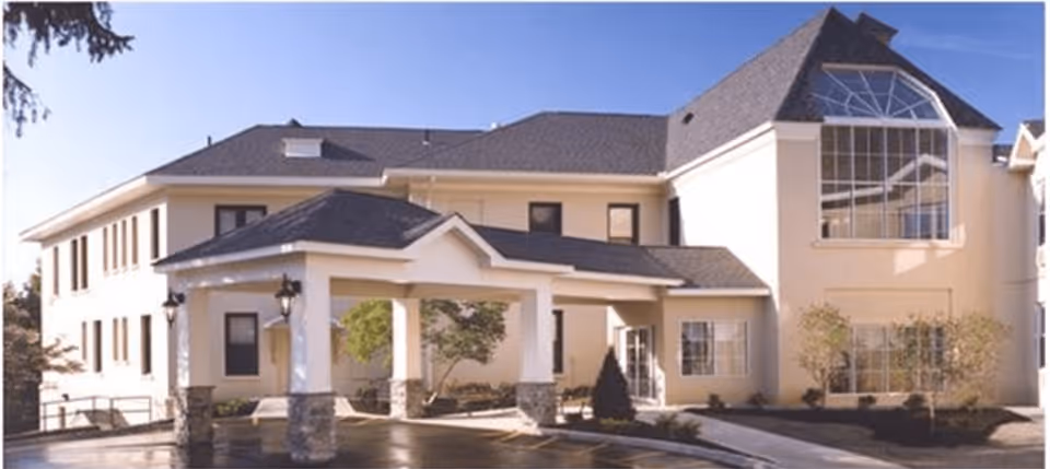 Exterior view of Promenade at Tuxedo Place, a multi-story senior living facility with a covered entrance supported by columns, large windows, and a well-maintained driveway and landscaping.