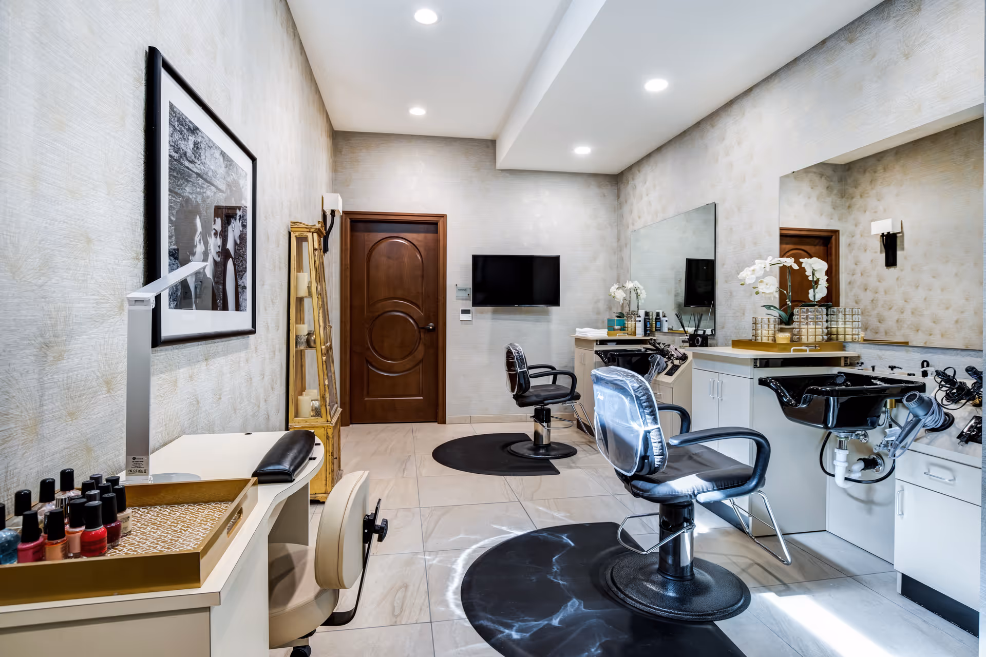 Interior view of a salon area in a senior living facility with two black salon chairs in front of sinks and mirrors, a manicure station with nail polish bottles, a wooden door, a wall-mounted TV, and decorative elements including a framed black and white photo and a tall mirror with candles.