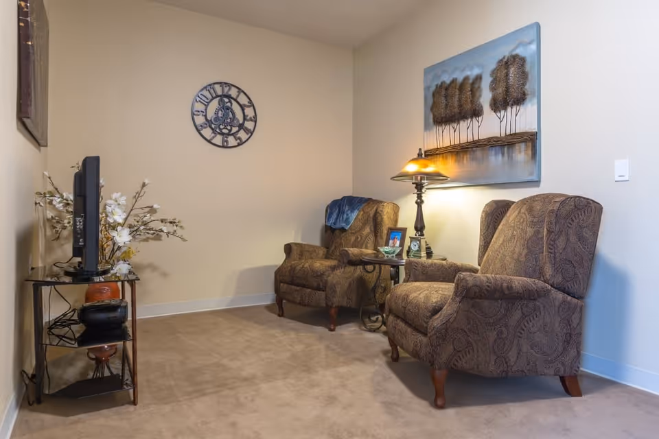 A cozy sitting area with two patterned armchairs separated by a small side table holding a lamp, a framed photo, and a clock. A wall clock and a painting of trees above a body of water hang on the beige walls. A small TV sits on a glass stand with decorative flowers beside it.