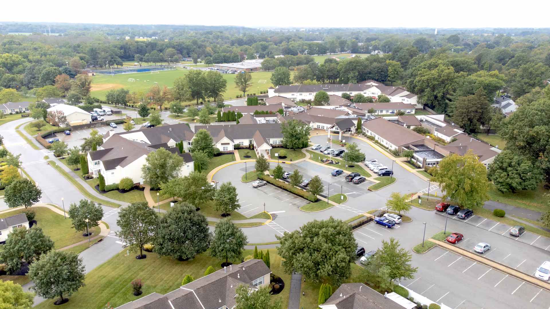 Aerial view of Friends Village senior living campus with multiple low-rise buildings, parking lots, driveways, and surrounding trees and lawns.