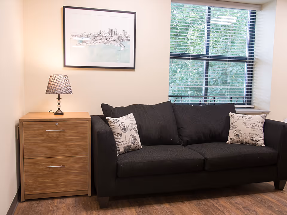 Black couch with patterned pillows next to a wooden side table and lamp beneath framed artwork and a window.