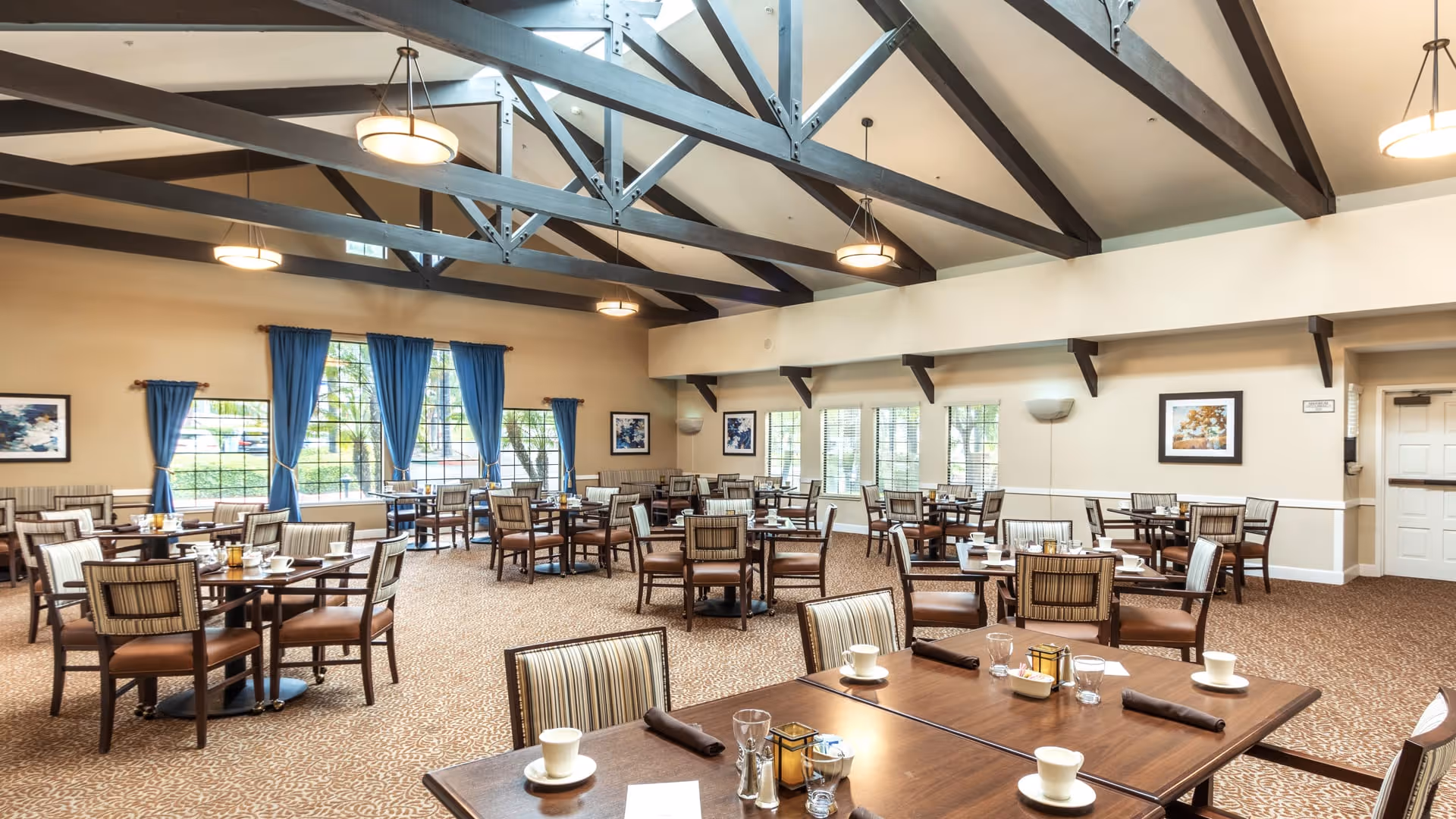 A spacious dining room with multiple wooden tables and chairs arranged neatly. Each table is set with cups, glasses, napkins, and condiments. The room features large windows with blue curtains, beige walls, and exposed dark wooden ceiling beams with hanging light fixtures. Framed artwork decorates the walls.