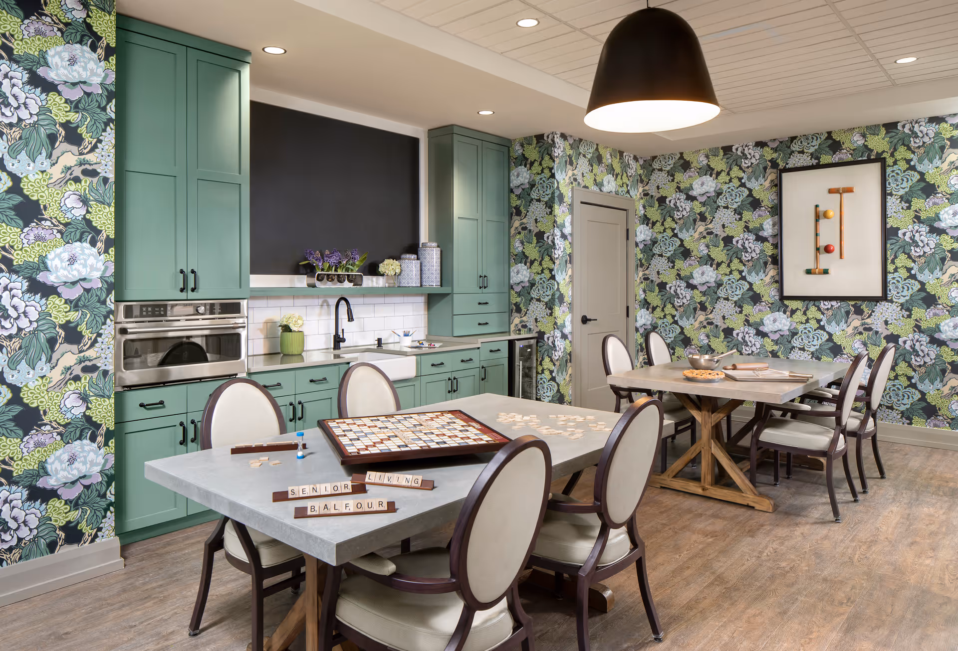 A bright and cozy common room with green cabinetry and floral wallpaper. The room features two tables with chairs around them, one table has a Scrabble game in progress with tiles spelling 'SENIOR LIVING BALFOUR'. The room has a modern farmhouse sink, built-in oven, and a large black pendant light hanging from the ceiling.