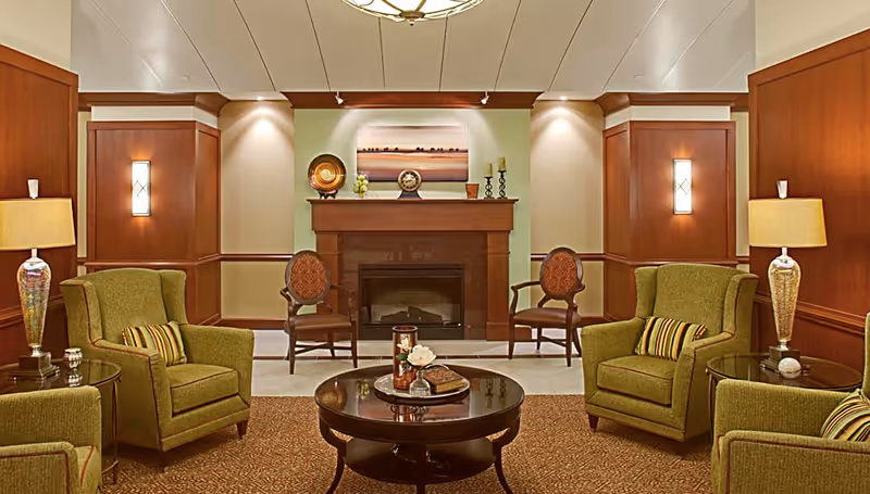 A cozy senior living room with green upholstered armchairs, a round wooden coffee table with decorative items, two wooden chairs with patterned cushions, a fireplace with a mantel decorated with candles and a clock, warm lighting from wall sconces and table lamps, and wood-paneled walls.