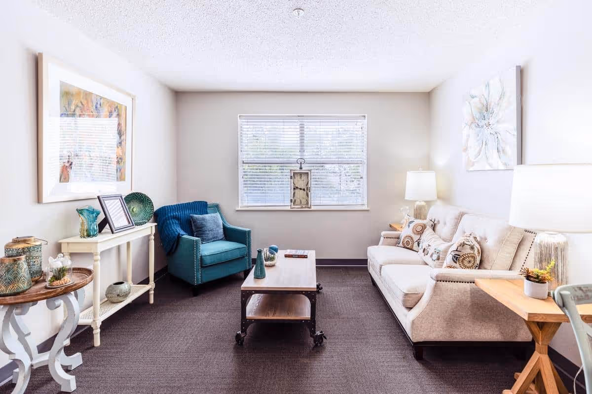 A bright and cozy living room with a beige sofa adorned with patterned cushions, a teal armchair with a blue throw blanket, a wooden coffee table with decorative items, and two side tables with lamps. The room has a large window with blinds, light-colored walls, and artwork hanging on the walls.