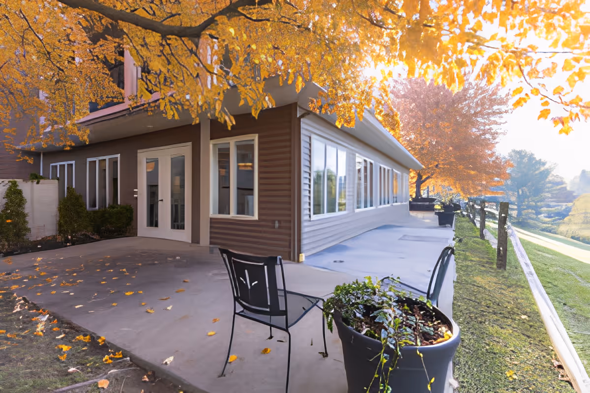 Outdoor patio area of a senior living facility with a concrete walkway, metal chairs, potted plants, and trees with yellow autumn leaves.