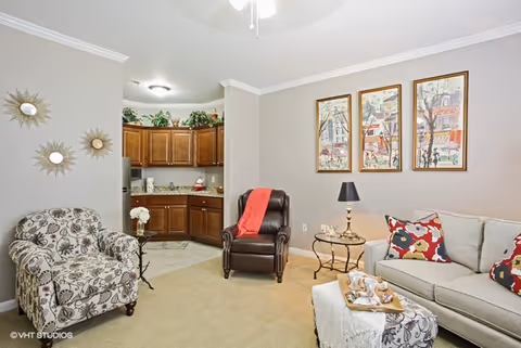 A cozy living room area with a beige carpet, a floral patterned armchair, a dark brown leather recliner with a red throw, and a light gray sofa with colorful floral pillows. There is a small round side table with a lamp and decorative items next to the recliner. Three framed artworks hang on the wall above the sofa. In the background, a kitchen with wooden cabinets and granite countertops is visible. The walls are painted light gray and the ceiling has crown molding and a ceiling fan.