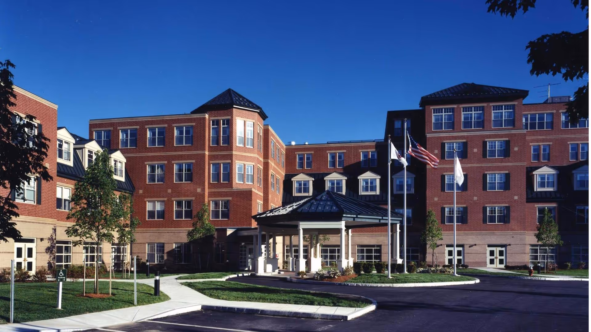 Front exterior of a multi-story red-brick assisted living building with a covered circular driveway, flagpoles, and landscaped grounds under a clear blue sky.