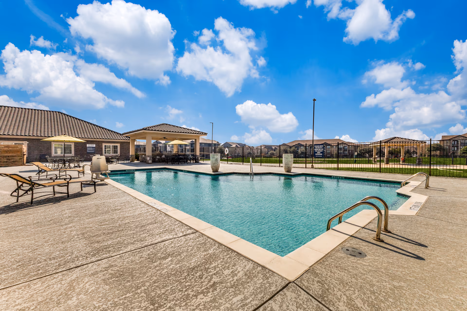 Outdoor swimming pool area at a senior living facility with lounge chairs, tables with umbrellas, and a covered seating area. The pool is surrounded by a concrete deck and a black metal fence. Residential buildings are visible in the background under a bright blue sky with scattered clouds.