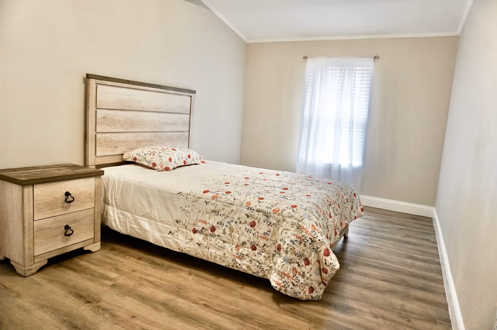 A simple bedroom with a wooden bed frame and matching nightstand. The bed is made with white sheets and a floral patterned comforter and pillow. There is a window with sheer white curtains allowing natural light into the room. The floor is wood laminate and the walls are painted a light beige color.