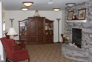 A cozy living room featuring a stone fireplace with a mantle decorated with framed pictures and small sculptures. To the left, there is a wooden entertainment center with shelves holding books and decorative items. Two red upholstered chairs and a small round wooden table are positioned near the entertainment center. The room has carpeted flooring and ceiling lights.