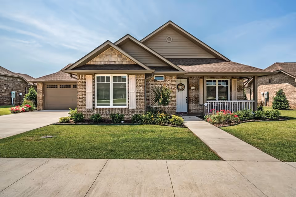 Single-story brick house with a front porch, attached garage and a landscaped lawn under a clear blue sky.