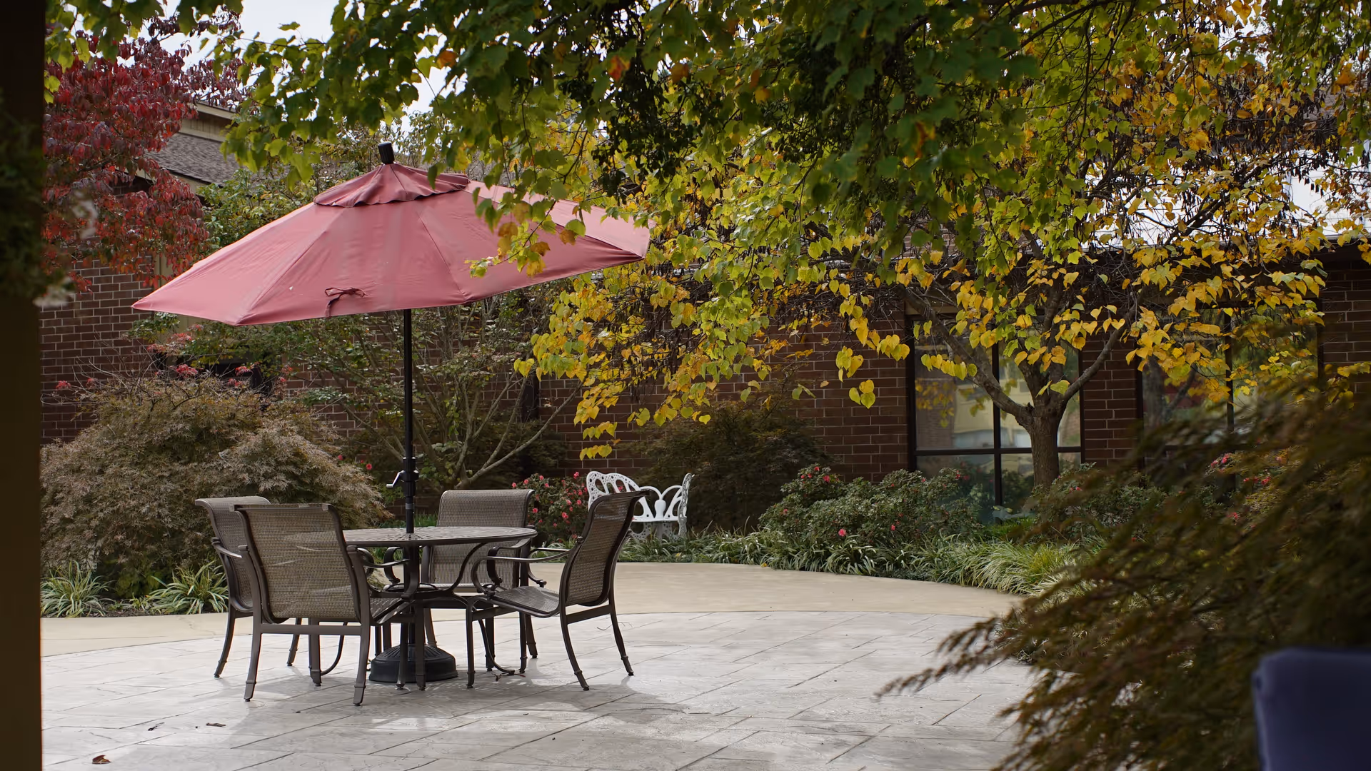 Outdoor patio area with a round table and four chairs under a large red umbrella, surrounded by trees with green and yellow leaves and a brick building in the background.