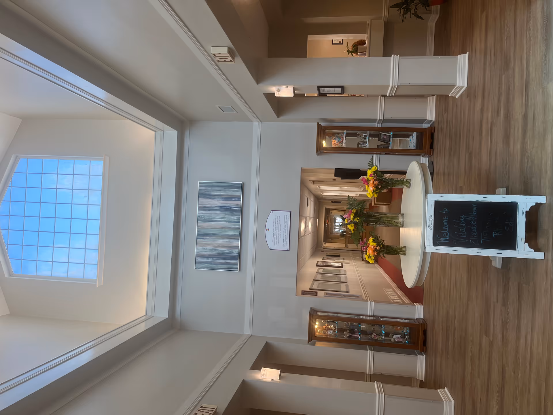 Bright senior living lobby interior with a round table of flower vases, a skylight, hallway and display cabinets.