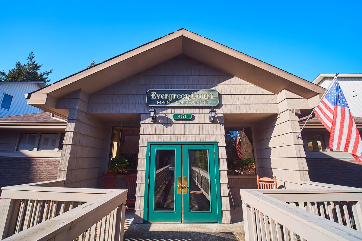Front entrance of a building with a peaked roof and green double doors. A sign above the doors reads 'Evergreen Court Main Entrance' with the number 451 below it. There is an American flag on the right side and a wooden ramp leading up to the entrance.