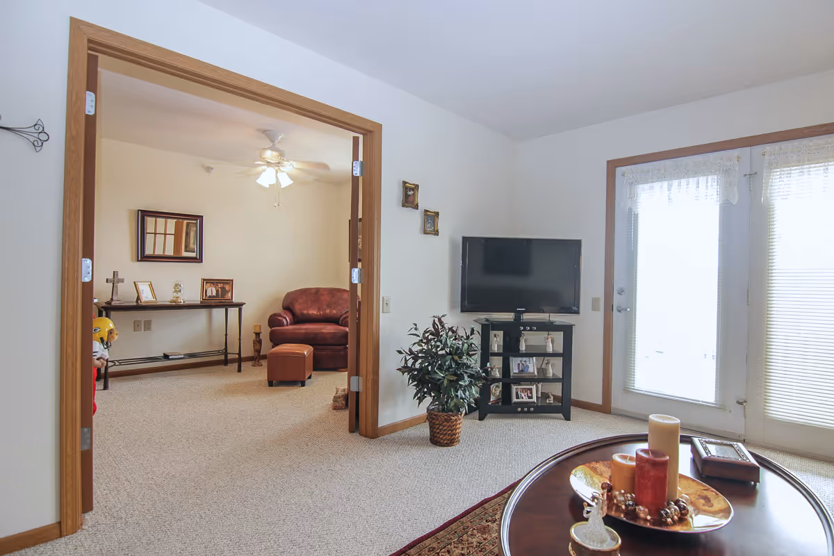 Interior view of a senior apartment living area with a TV on a stand, a potted plant, and a round coffee table with candles and decorative items. Through an open doorway, there is another room with a brown leather armchair, ottoman, a console table with framed pictures, and a ceiling fan with lights.