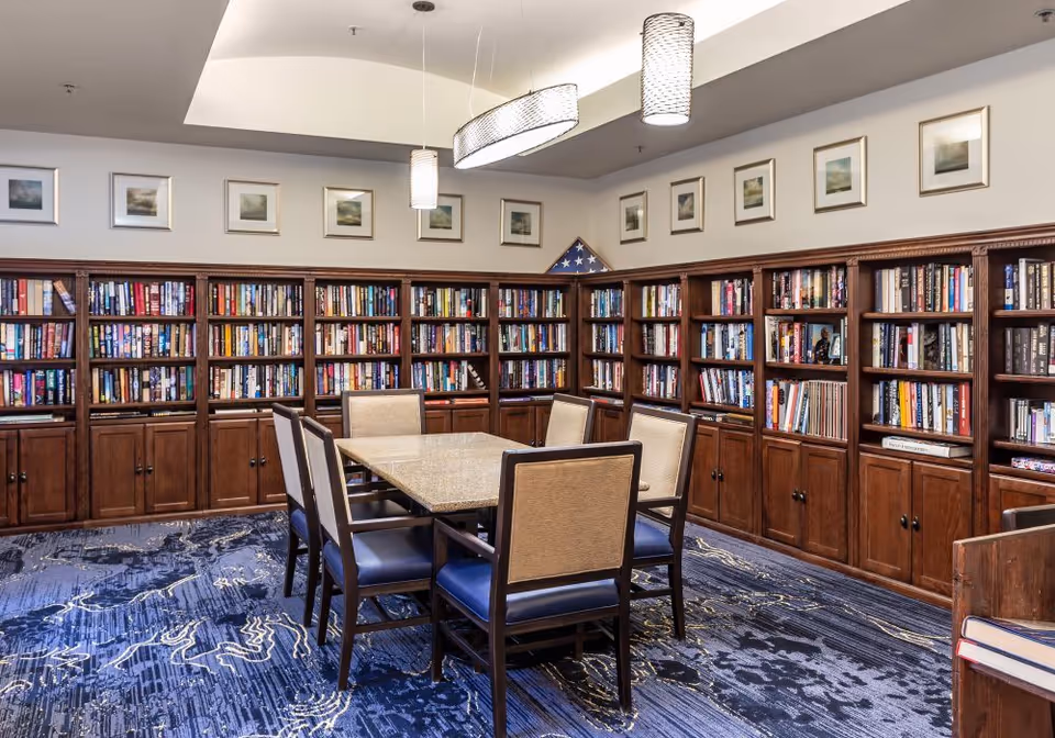 A cozy library room with wooden bookshelves filled with books lining the walls. A rectangular table with six chairs is placed in the center of the room. The ceiling has modern hanging light fixtures, and framed pictures are displayed above the bookshelves. The carpet has a blue and black abstract pattern.
