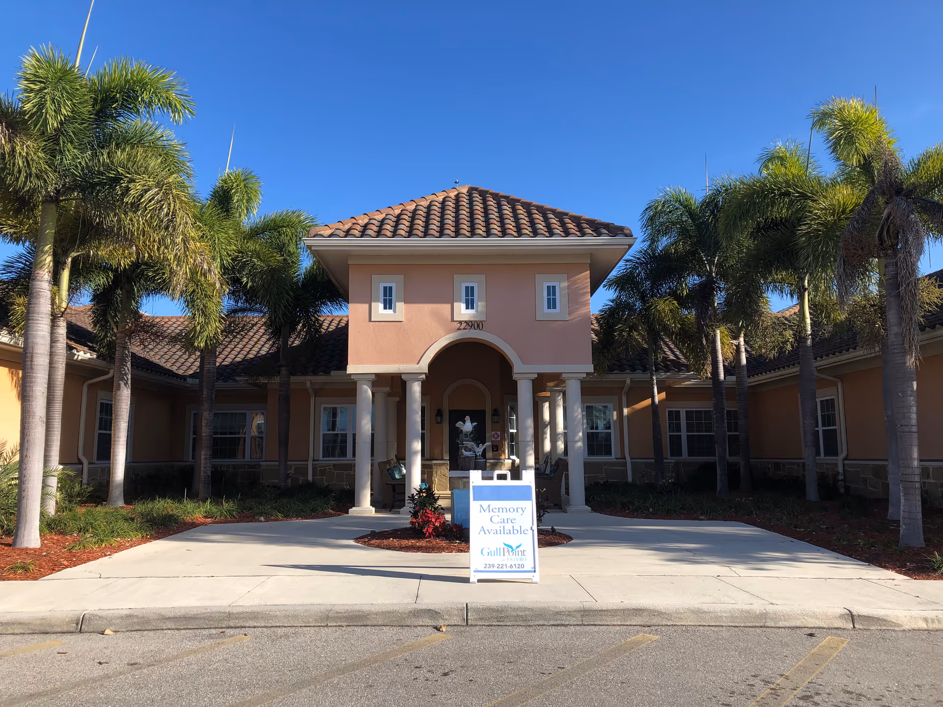 Front entrance of the Gulf Coast Memory Care building with palm trees and a 'Memory Care Available' sign on the driveway.