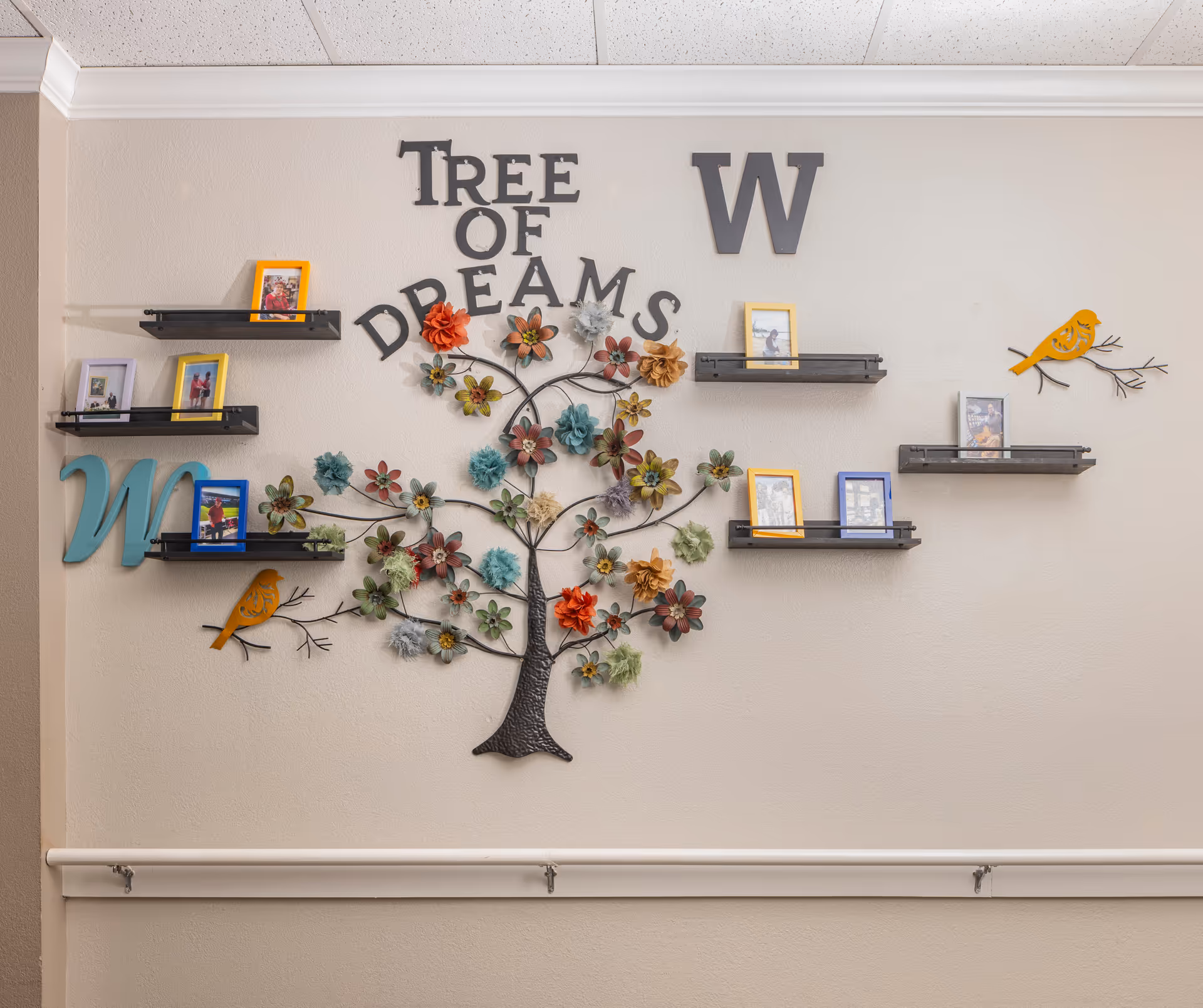 Wall decorated with a metal art piece shaped like a tree with colorful flowers and birds, surrounded by small shelves holding framed photos. The words 'TREE OF DREAMS' and large letters 'W' and 'M' are displayed on the wall.
