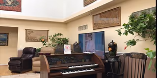 Interior view of a living room area with a piano in the foreground, a flat-screen TV mounted on the wall, comfortable seating including a leather recliner and a beige sofa, decorative plants, and framed artwork on the walls.