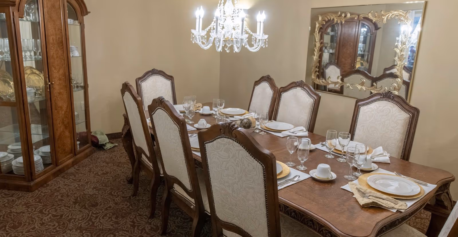 A formal dining room with a long wooden table set for eight people. The table is arranged with white plates, gold chargers, glassware, and folded napkins. There are eight upholstered chairs with wooden frames around the table. A chandelier with lit bulbs hangs above the table. A large ornate mirror is mounted on the wall, reflecting a wooden china cabinet filled with dishes and glassware. The room has beige walls and patterned carpet flooring.