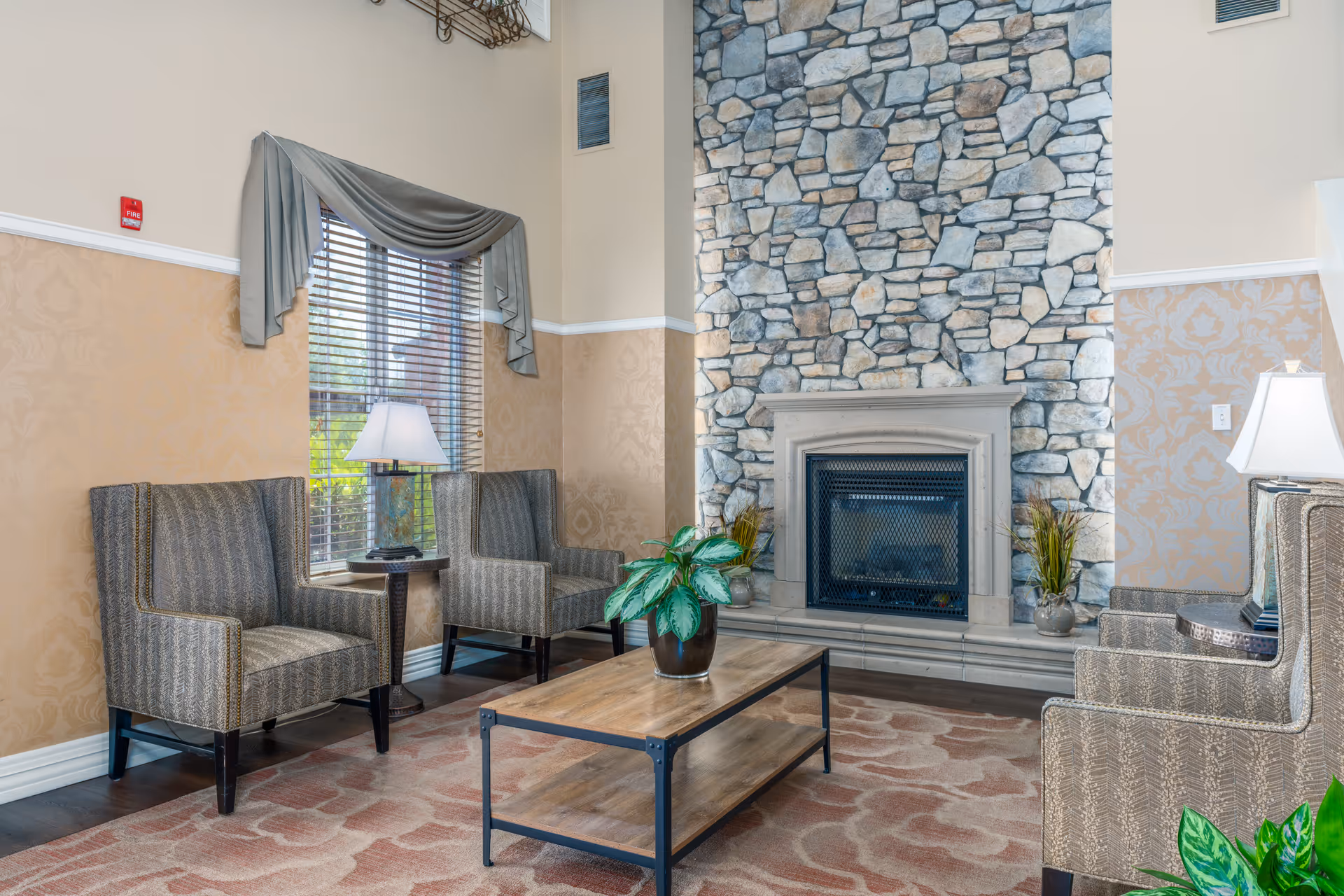 A cozy sitting area with four patterned armchairs arranged around a wooden coffee table with a potted plant. The room features a tall stone fireplace, beige walls with a decorative pattern, a window with blinds and a gray valance, and two table lamps on side tables beside the chairs.