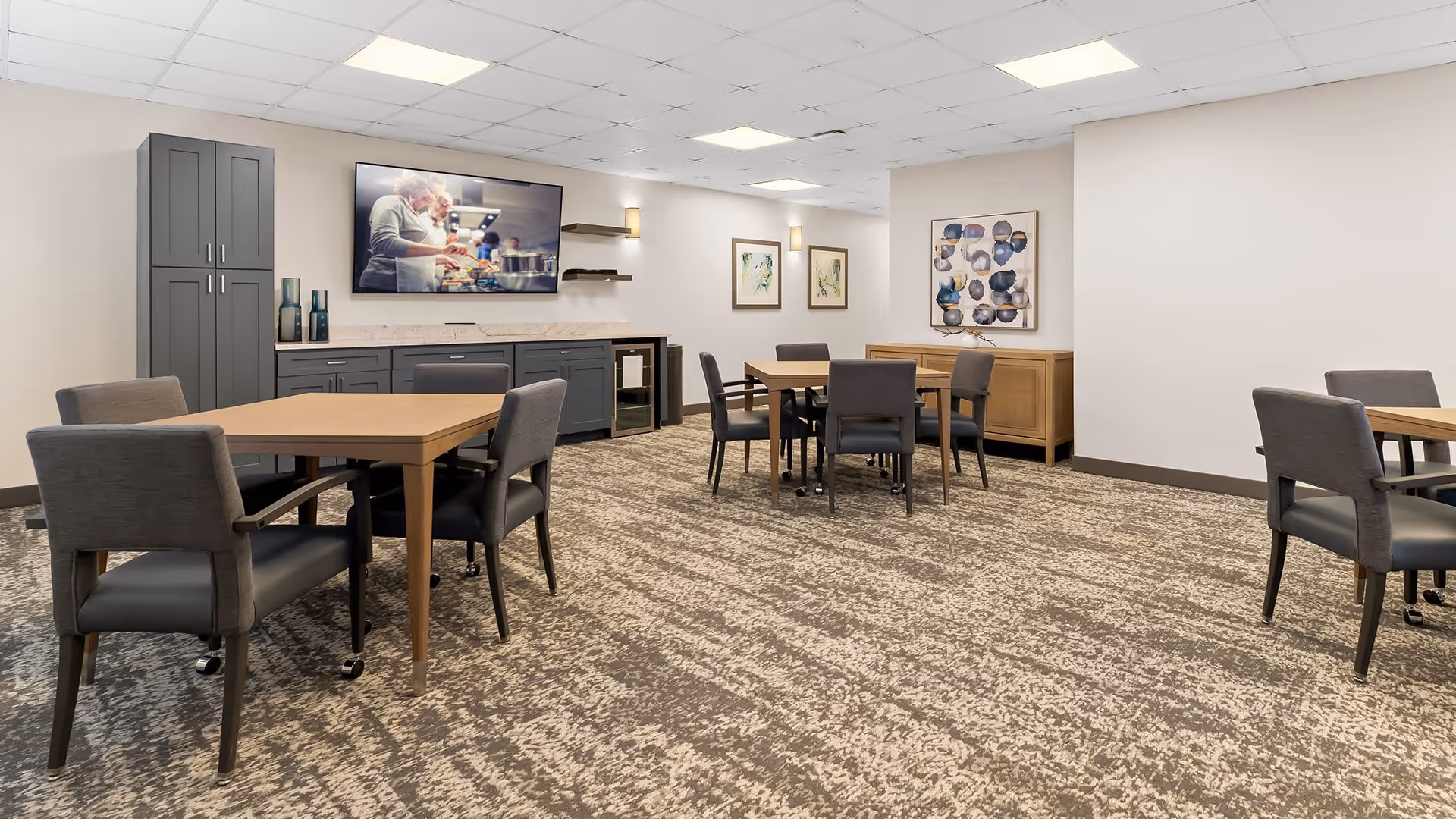 A modern dining room with multiple wooden tables and gray upholstered chairs on a patterned carpet. The room features a gray cabinet and countertop along one wall with decorative vases and a flat-screen TV mounted above showing an elderly couple cooking. The walls are decorated with framed abstract artwork, and the ceiling has recessed lighting.