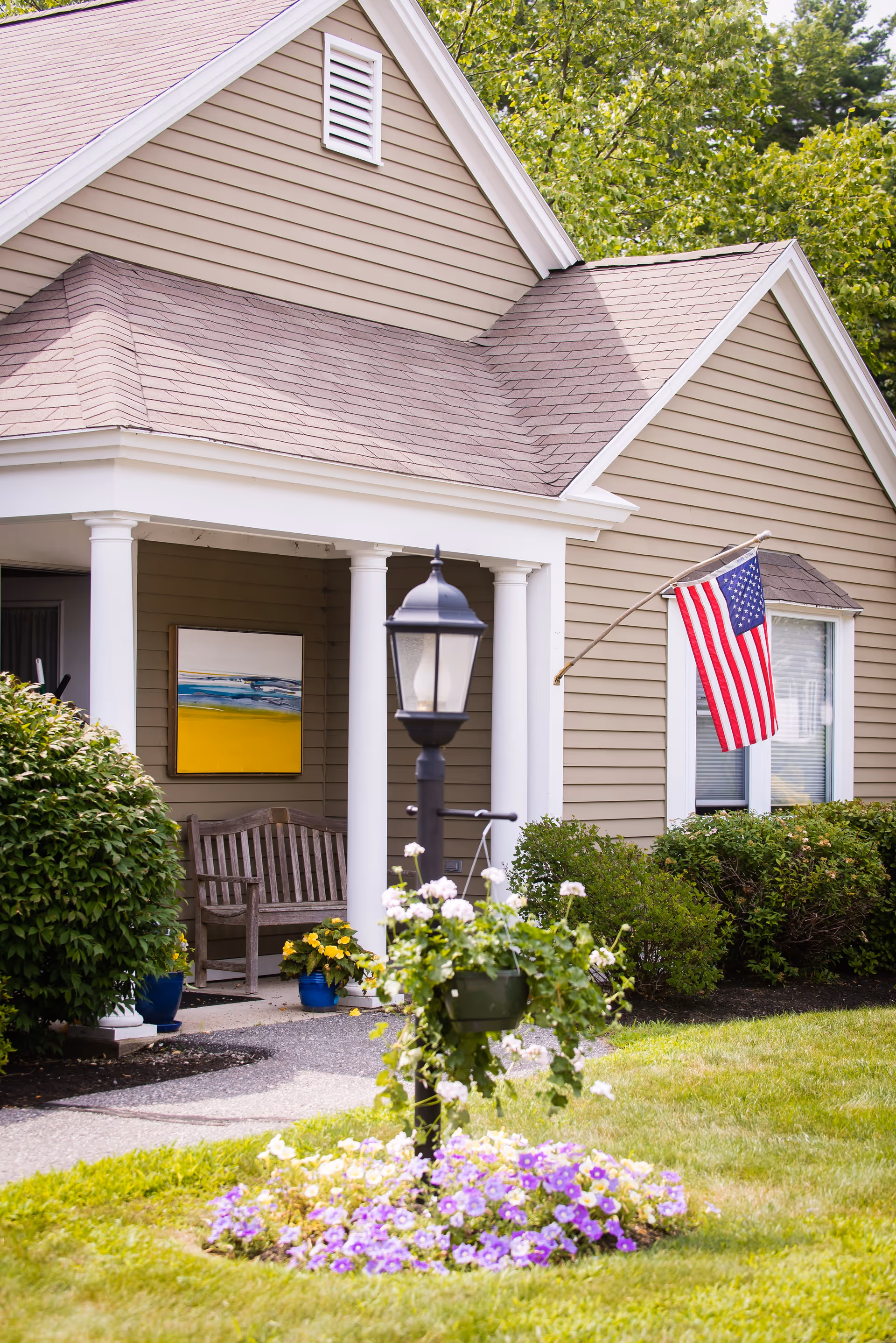 Exterior view of a beige house with white trim, featuring a small porch with white columns, a wooden bench, potted plants, an American flag mounted on the wall, a black lamp post with hanging flower baskets, and a flower bed with purple and white flowers in the foreground.