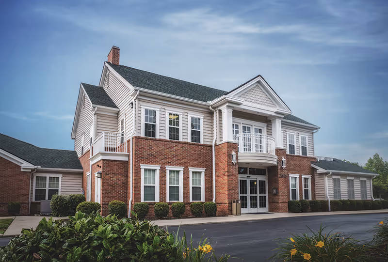 Exterior view of a two-story brick and siding building with white columns and multiple windows under a blue sky, surrounded by bushes and a paved driveway.