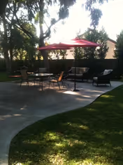 Outdoor patio area with a red umbrella shading a table surrounded by chairs and cushioned seating, set on a concrete surface with grass and trees in the background.