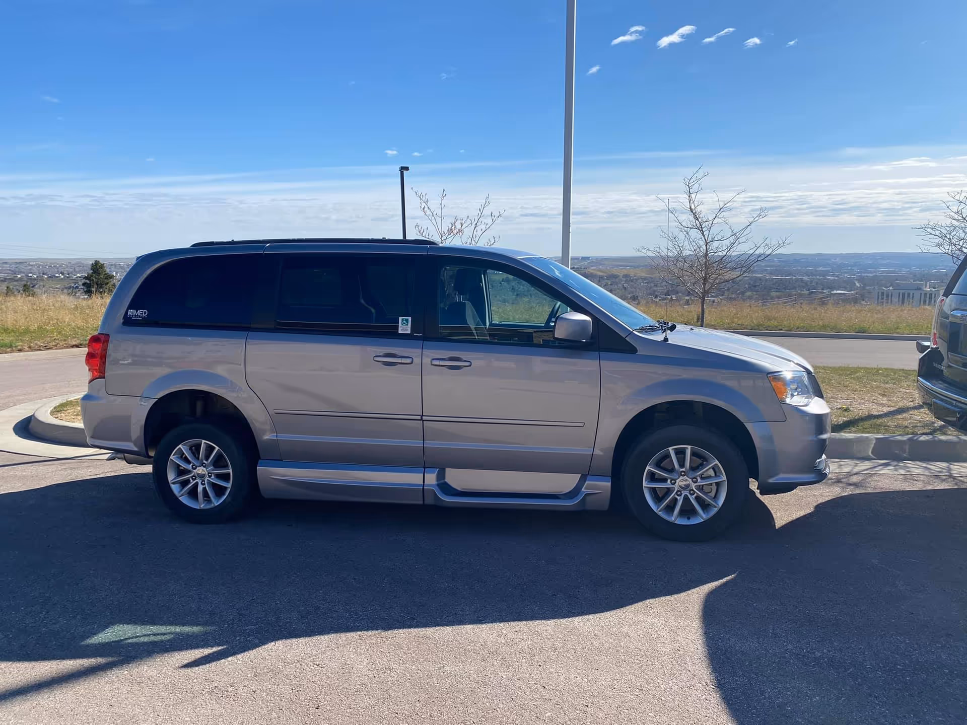 A silver minivan parked in an outdoor parking lot with a clear blue sky and distant landscape in the background. The minivan has a handicap sticker on the window and a ramp on the side, indicating it is adapted for wheelchair access.