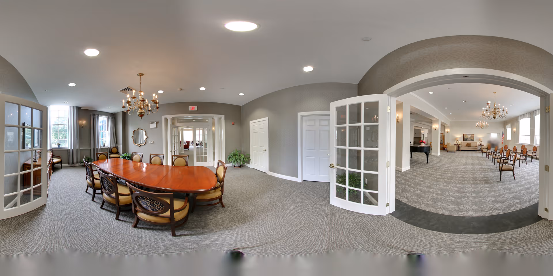 Panoramic interior of a senior living community room featuring a long wooden dining/meeting table with upholstered chairs, chandeliers, and an adjacent large multipurpose seating area visible through French doors.