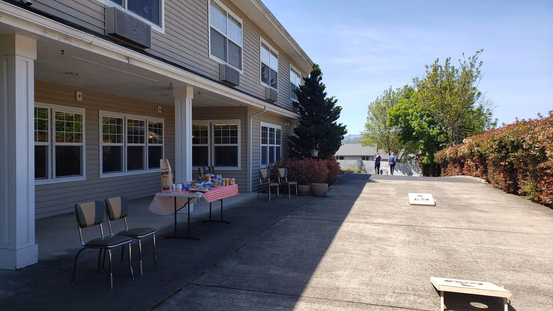 Outdoor patio area of a senior living facility with a table covered in a red and white checkered tablecloth holding snacks and drinks. Several chairs are arranged nearby. Two people are walking in the distance along a concrete pathway lined with bushes and trees under a clear blue sky.