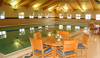 Indoor swimming pool area with wooden ceiling beams and multiple windows. There are wooden tables and chairs with blue cushions arranged near the poolside, and a small floral centerpiece on one of the tables.