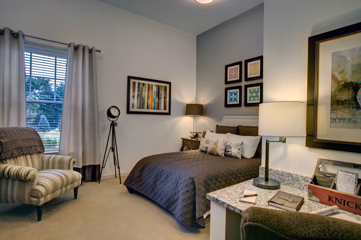 A cozy bedroom featuring a bed with a dark quilted bedspread and multiple decorative pillows. The room has a striped armchair with a throw blanket near a window with curtains. There is a tripod floor clock, a nightstand with a lamp, and framed artwork on the walls. A granite countertop with a table lamp, notebook, and a wooden box labeled 'KNICK' is visible in the foreground.