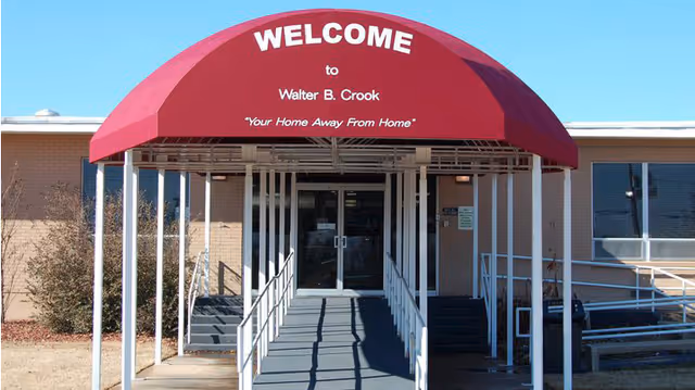 Entrance of Walter B. Crook Nursing Facility with a red canopy that reads 'WELCOME to Walter B. Crook Your Home Away From Home'. There is a ramp and stairs leading to glass double doors of the building.