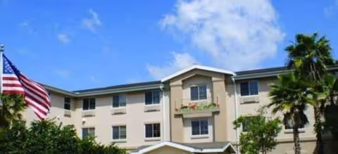 Three-story beige senior living building exterior with flag and palm trees against blue sky.