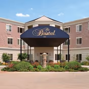 Front exterior view of The Bristal Assisted Living at Armonk building with a covered entrance canopy displaying the facility name, surrounded by landscaped greenery and a clear blue sky.