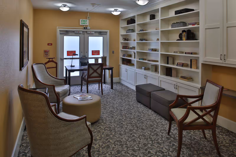 A cozy interior room with patterned carpet, several upholstered chairs, a small round ottoman, and a wooden desk with two chairs near a set of double exit doors. One wall features built-in white shelves filled with books and decorative items, and there are two gray cushioned benches below the shelves.
