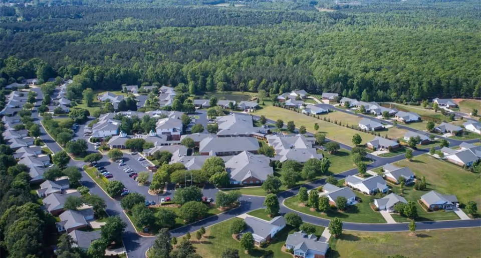 Aerial view of Summit Hills senior living facility showing multiple single-story buildings with gray roofs arranged along curved roads, surrounded by green lawns and trees, with a dense forest in the background.